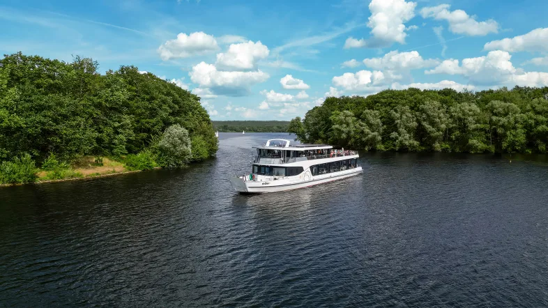 Foto eines Fahrgastschiffes mit zwei Decks, auf denen viele Touristen die Rundfahrt auf dem Halterner Stausee genießen. Links und rechts neben dem Schiff sind grün bewachsene Inseln zu sehen und die Schneise, durch die das Schiff durchgefahren ist. Der Himmel ist blau und mit einigen weißen Wolken durchzogen, das Wasser spiegelt leicht den Himmel und das Schiff wider.