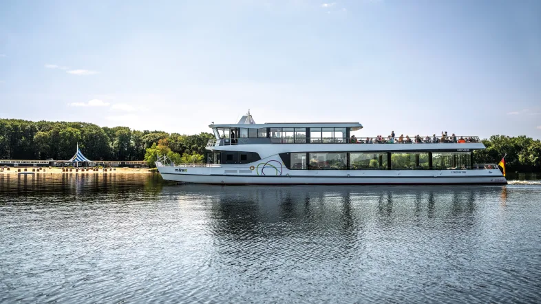 Zu sehen ist das Fahrgastschiff Möwe, während es auf seiner Rundfahrt einen bestimmten Teil des Halterner Stausees erreicht. Das Schiff besteht aus einem Außendeck, auf dem einige Passagiere das Wetter genießen und einem Unterdeck, das verglaste Scheiben aufweist. Das Wetter ist super, die Sonne scheint vom strahlend blauen Himmel. Im Hintergrund sieht man das Seebad in Haltern, an einem Sandstrand stehen einige Strandkörbe und ein riesiges Sonnensegel.