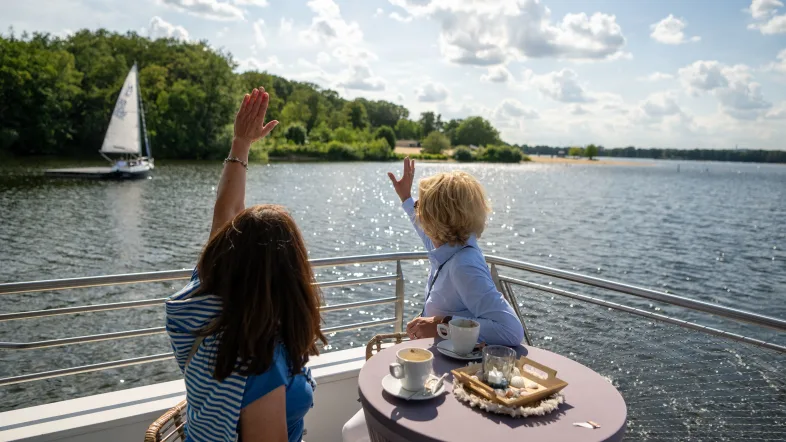 Zwei Frauen sitzen auf der hinteren Terrasse des Ausflugsschiffs und haben draußen in der Sonne auf einem Tisch zwei Cappuccino Tassen vor sich stehen. Sie sitzen dem Segelboot zugewandt, das sich weiter links im Bild befindet. Beide begrüßen den Segler mit winkenden Armen.