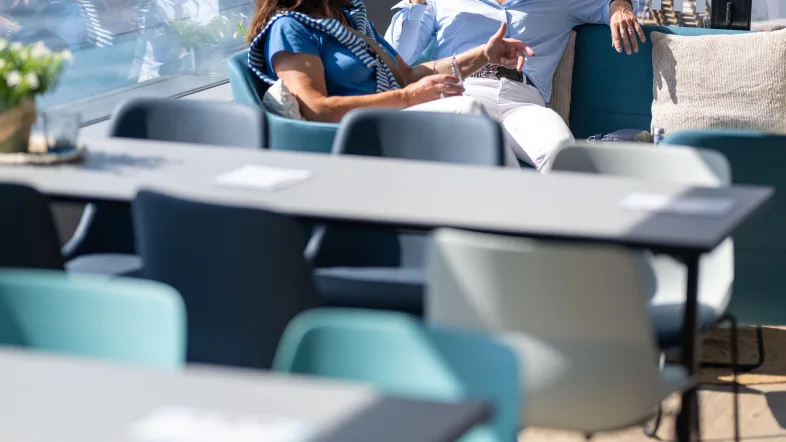 Das Bild zeigt zwei Damen, die sich auf dem Unterdeck des Schiffes in einer möblierten Lounge austauschen und dabei ein kaltes Getränk zu sich nehmen. Im Vordergrund wird die Bestuhlung des Unterdecks gezeigt. Auf einem warmen Holzboden finden sich graue Tische, an denen maximal 6 Personen sitzen können. Die Stühle weisen verschiedene Farben auf, von hellblau bis türkis gibt es sogar gelbe Stühle. Die Sonne scheint in das Schiff hinein und erzeugt eine sommerliche Atmosphäre.
