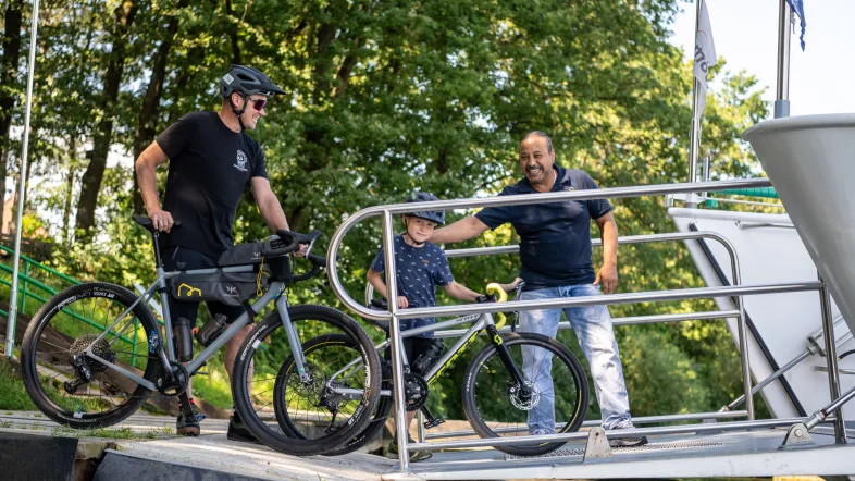 Das Bild zeigt den Bug des Schiffes, über den ein barrierefreier Zugang zum Ausflugsschiff erfolgen kann. Hier zu sehen ist der Bootsmann, der die Gäste in Empfang nimmt. Ein Mann mittleren alters und ein Kind, ca.10 Jahre alt, laufen gemeinsam mit ihren Fahrrädern auf die Möwe.
