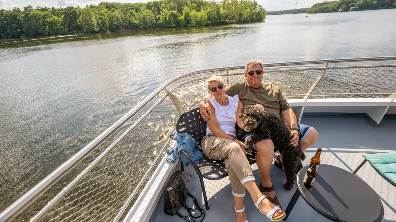 Ein glückliches Pärchen sitzt entspannt auf dem Sonnendeck der Möwe, gemeinsam mit ihrem schwarzen, lockigen Hund. Beide tragen Sonnenbrillen und lächeln in die Kamera. Die Frau lehnt sich liebevoll an den Mann, der den Hund halb auf dem Schoß hält. 
Neben ihnen stehen ein kleiner runder Tisch mit Kaltgetränken. Im Hintergrund sieht man den Halterner Stausee und leicht bewölkten Himmel.