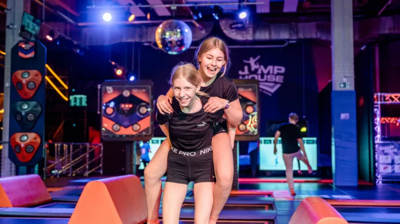 Zwei Mädchen stehen lachend und Huckepack vor einer Trampolinfläche