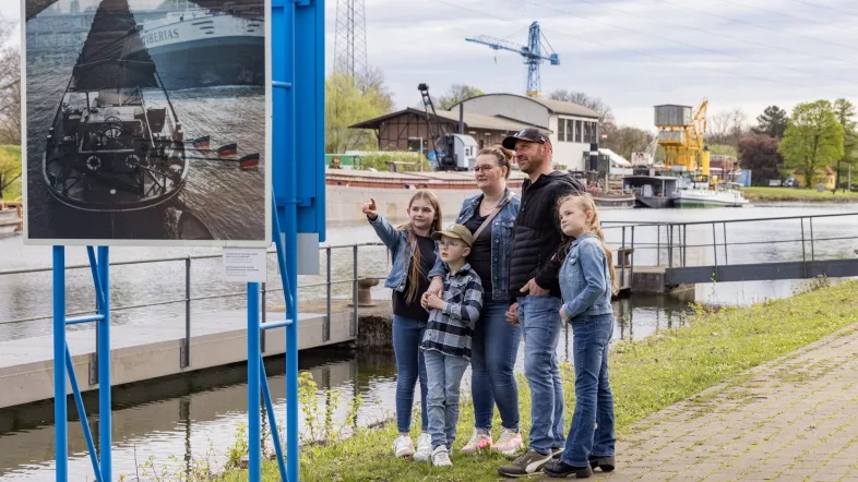 Eine Familie sieht sich eine Ausstellungstafel am Ufer des Oberwassers an.