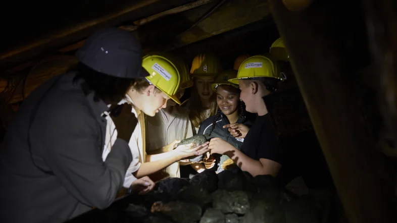 Eine Gruppe von Schüler:innen mit gelben Helmen im Stollen. Sie greifen ein Stück Kohle aus einem Förderwagen.