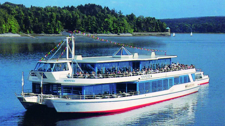 Idyllisches Postkartenmotiv: Fahrgastschiff auf dem Möhnesee mit spiegelndem Wasser und bewaldeten Ufern im Hintergrund.