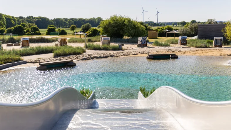 Die silberne Wellenrutsche führt ins glasklare Wasser des Nicht-Schwimmerbereichs im Naturbad Olfen. Im Hintergrund erkennt man den Sandstrand mit Strandkörben.