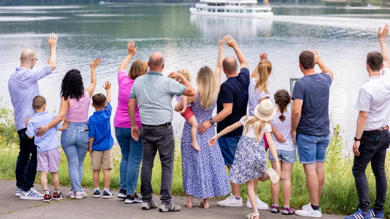 Menschen am Ufer des Biggesees winken einem vorbeifahrenden Ausflugsboot zu.