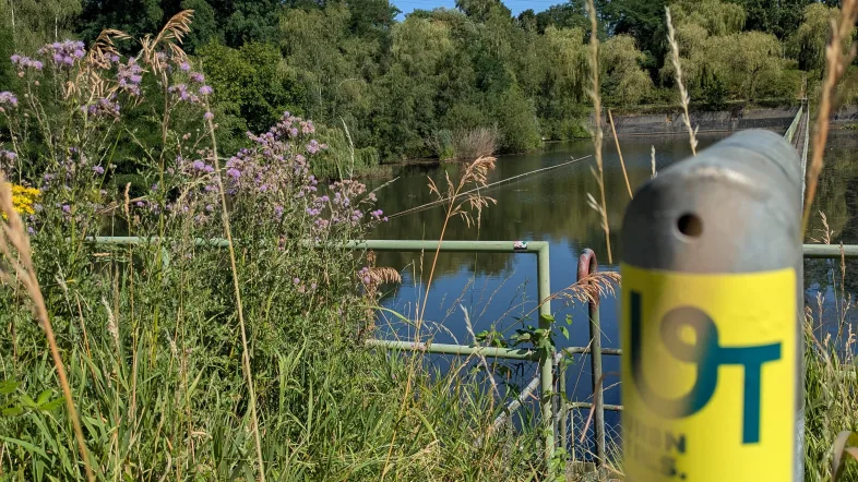Foto von einem Pfahl mit dem Urban.Trails Aufkleber an einem Gewässer.
