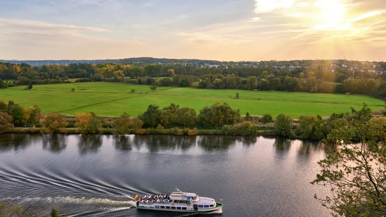Panoramablick auf die „Weiße Flotte“ Mülheim mit einem Ausflugsschiff auf dem Fluss und der Stadt im Hintergrund.
