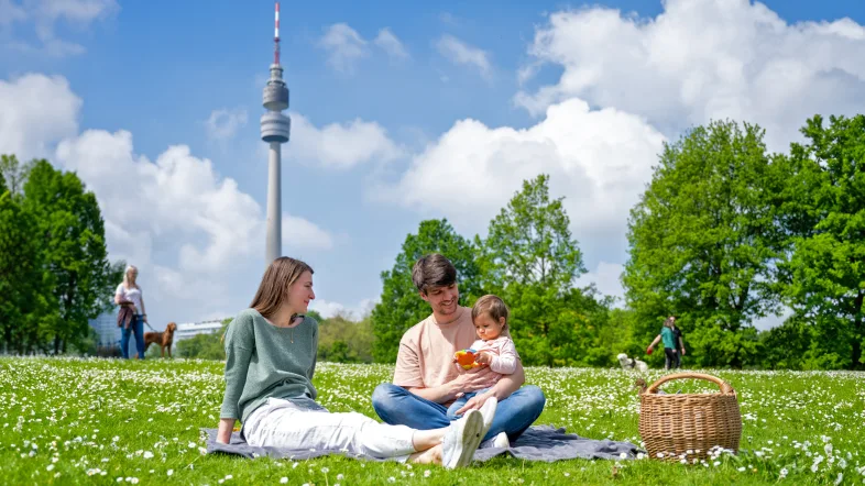 Familie beim Picknick auf einer blühenden Wiese im Westfalenpark Dortmund. Im Hintergrund der Florianturm, blauer Himmel mit weißen Wolken, umgeben von grünen Bäumen.