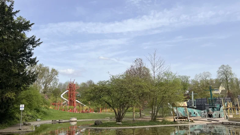 Blick auf den Robinson-Spielplatz im Westfalenpark Dortmund mit großem Kletterturm in Rot und langer Röhrenrutsche. Rechts im Bild ein Piratenschiff-Spielgerät, vorne ein Teich mit Spiegelung der Bäume.