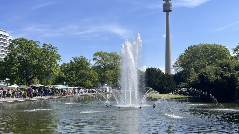 Springbrunnen im Flamingoteich im Westfalenpark Dortmund bei strahlendem Sonnenschein. Im Hintergrund ist der Florianturm zu sehen, links im Bild zahlreiche Besucher*innen entlang eines Markt- oder Veranstaltungsbereichs.