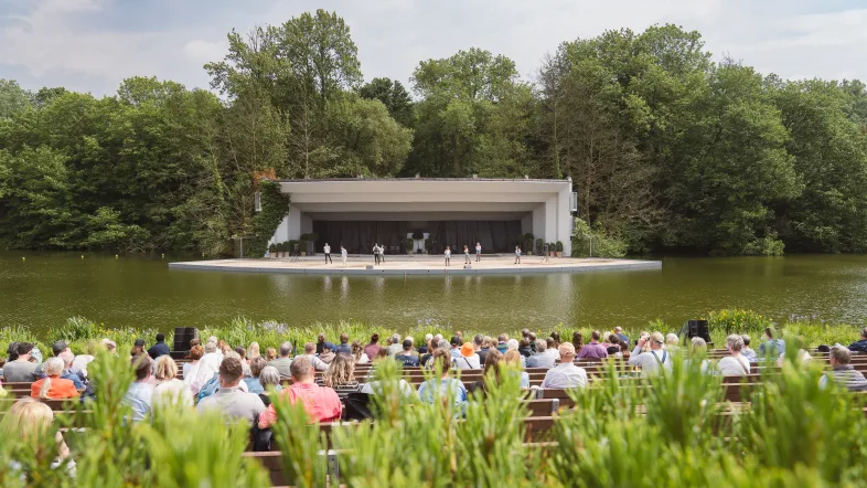 Publikum sitzt auf Bänken vor der Seebühne im Westfalenpark Dortmund. Die Bühne befindet sich auf dem Wasser, im Hintergrund dichte grüne Bäume. Auf der Bühne stehen mehrere Personen, offenbar bei einer Aufführung oder Begrüßung.