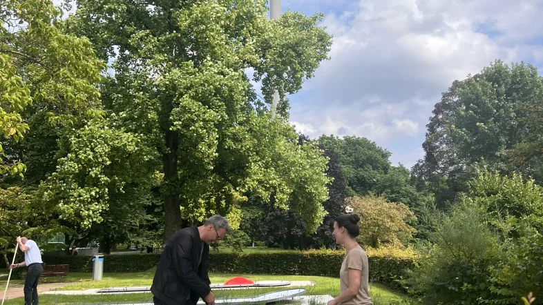Zwei Personen spielen Minigolf im Westfalenpark Dortmund auf einer der Betonbahnen. Im Hintergrund ist der Florianturm zwischen den grünen Bäumen sichtbar, darüber blauer Himmel mit Wolken.