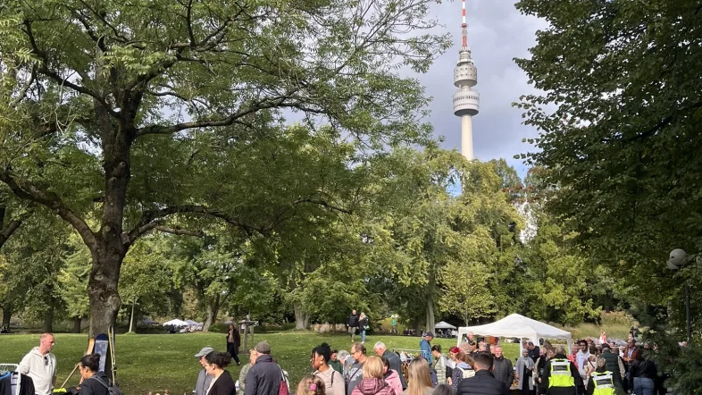 Zahlreiche Besucher*innen schlendern über den Trödelmarkt im Westfalenpark Dortmund. Zwischen den Bäumen sind Verkaufsstände mit Second-Hand-Waren aufgebaut, im Hintergrund ragt der Florianturm über das grüne Blätterdach hinaus.