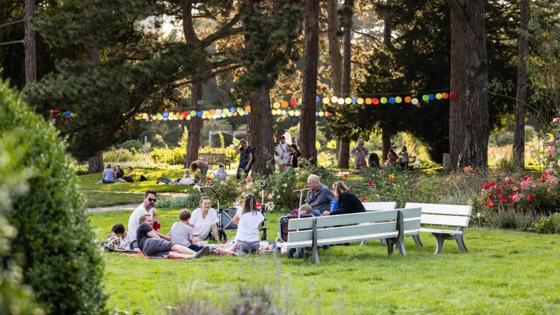 Menschen genießen einen sommerlichen Tag im Westfalenpark Dortmund: Familien und Gruppen sitzen auf Picknickdecken und Bänken zwischen Blumenbeeten und Bäumen. Bunte Lampions hängen zwischen den Bäumen und schaffen eine festliche Atmosphäre.