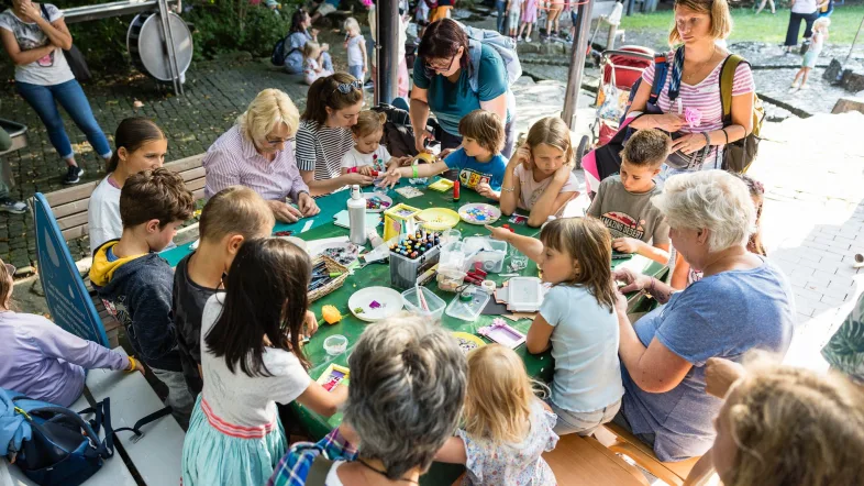 Kinder und Erwachsene sitzen gemeinsam an einem Basteltisch im Westfalenpark Dortmund. Auf dem Tisch liegen verschiedene Bastelmaterialien wie Stifte, Papier und Glitzersteine. Die Gruppe ist konzentriert bei der kreativen Arbeit, im Hintergrund sind weitere Parkbesucher*innen zu sehen.